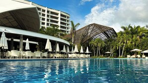 Ein Pool mit Liegestühlen und Sonnenschirmen, umgeben von Palmen und einem modernen Gebäude mit gewölbtem Dach.