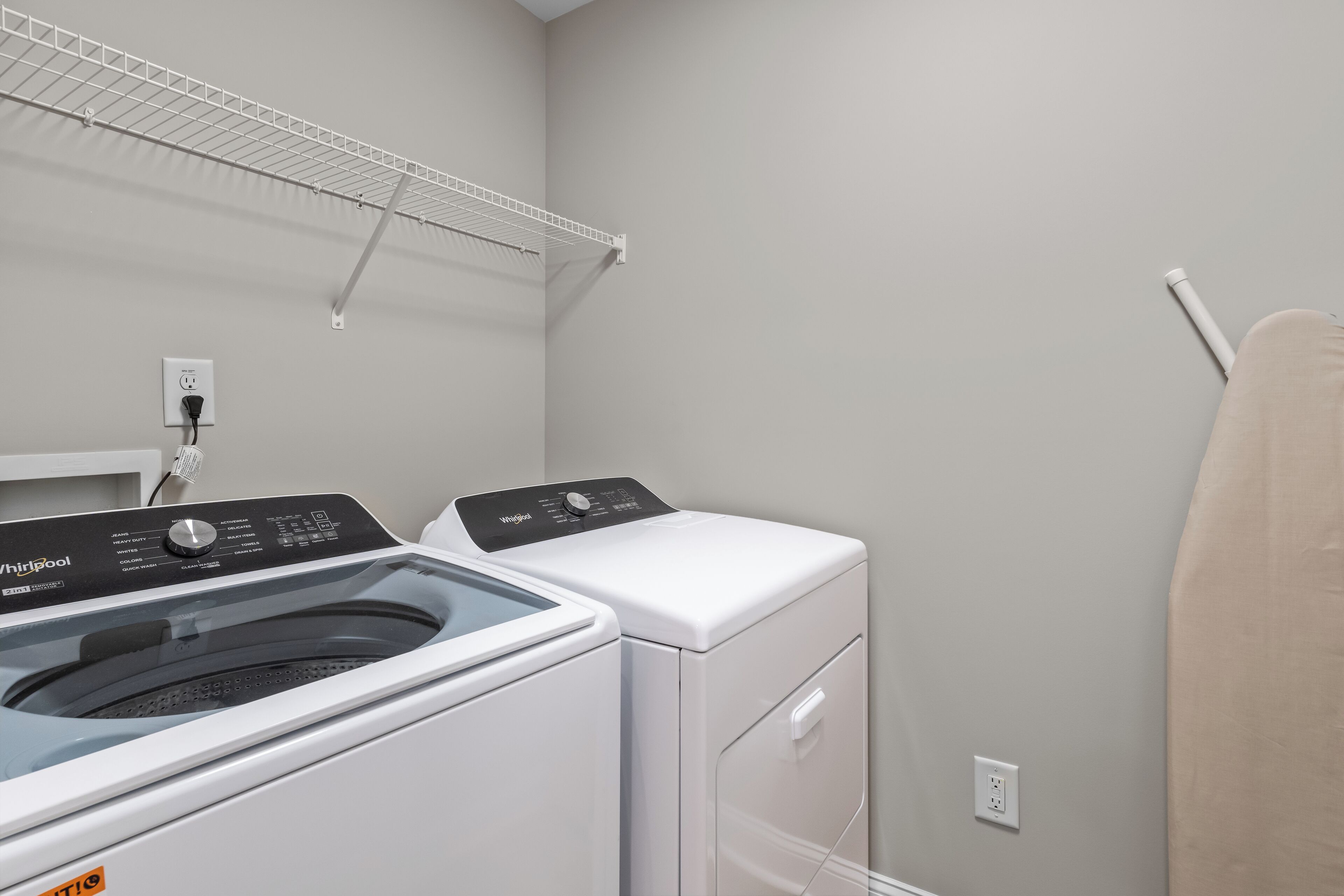 Un espace buanderie équipé d’un lave-linge et d’un sèche-linge à chargement frontal, d’une prise de courant et d’une étagère en métal située au-dessus du lave-linge.