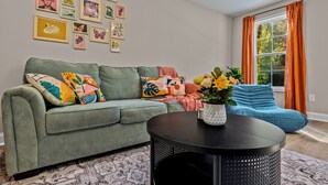 A living room with a green sofa, a black coffee table, and a blue bean bag. There are multiple framed artworks on the wall. - The Aspen ~ New, Trendy 3 Bed Near The Action (Kannapolis)