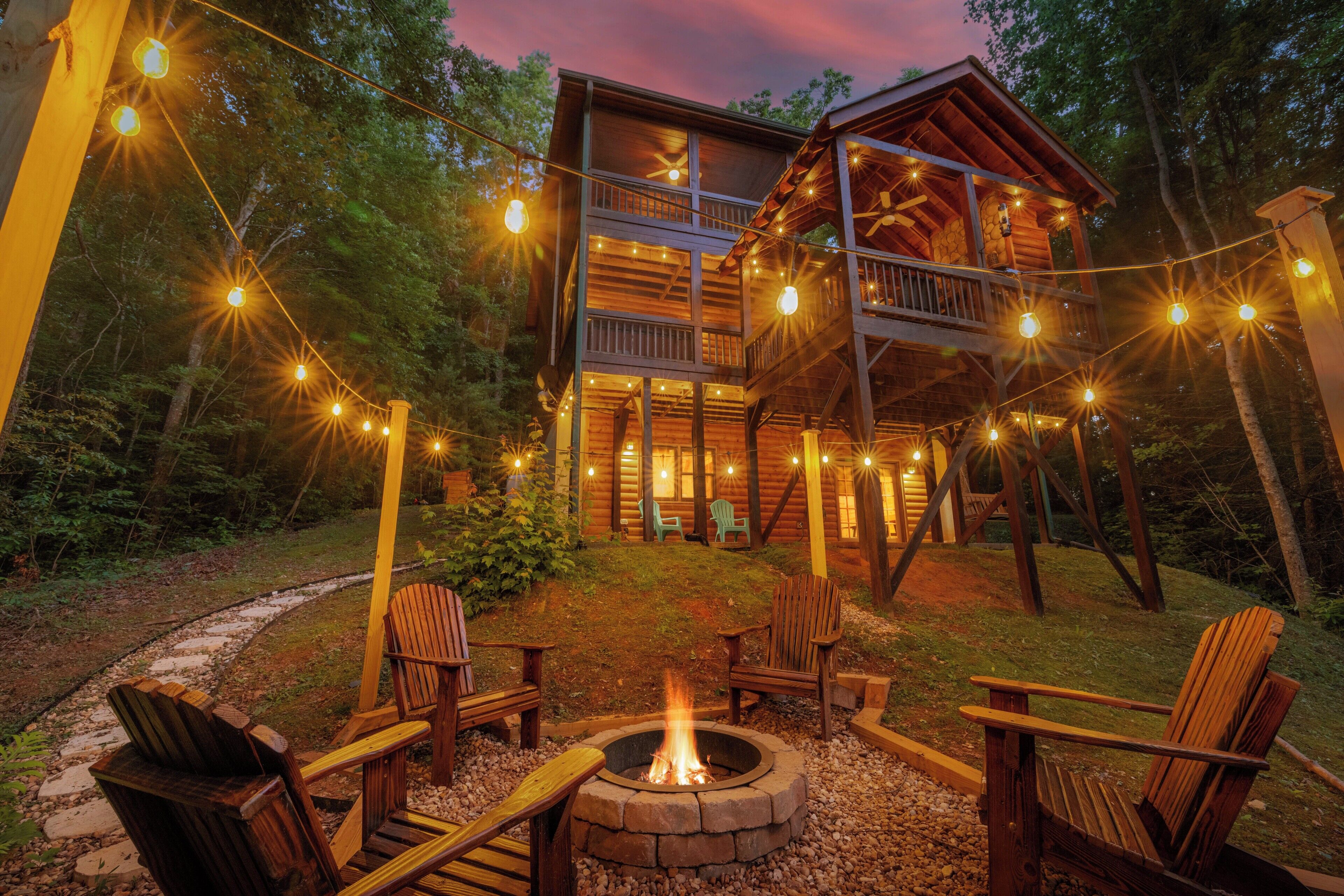 A wooden cabin with a deck, surrounded by trees, and a fire pit with chairs.
