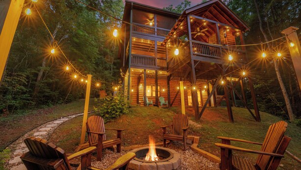 Eine Holzblockhütte mit einer Terrasse, umgeben von Bäumen, und einer Feuerstelle mit Sitzgelegenheiten.