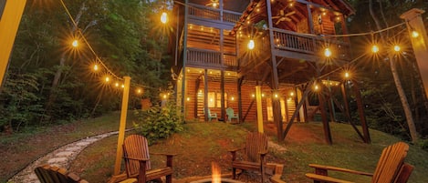 Une cabane en bois dotée d’une terrasse, entourée d’arbres, et un foyer avec des chaises.