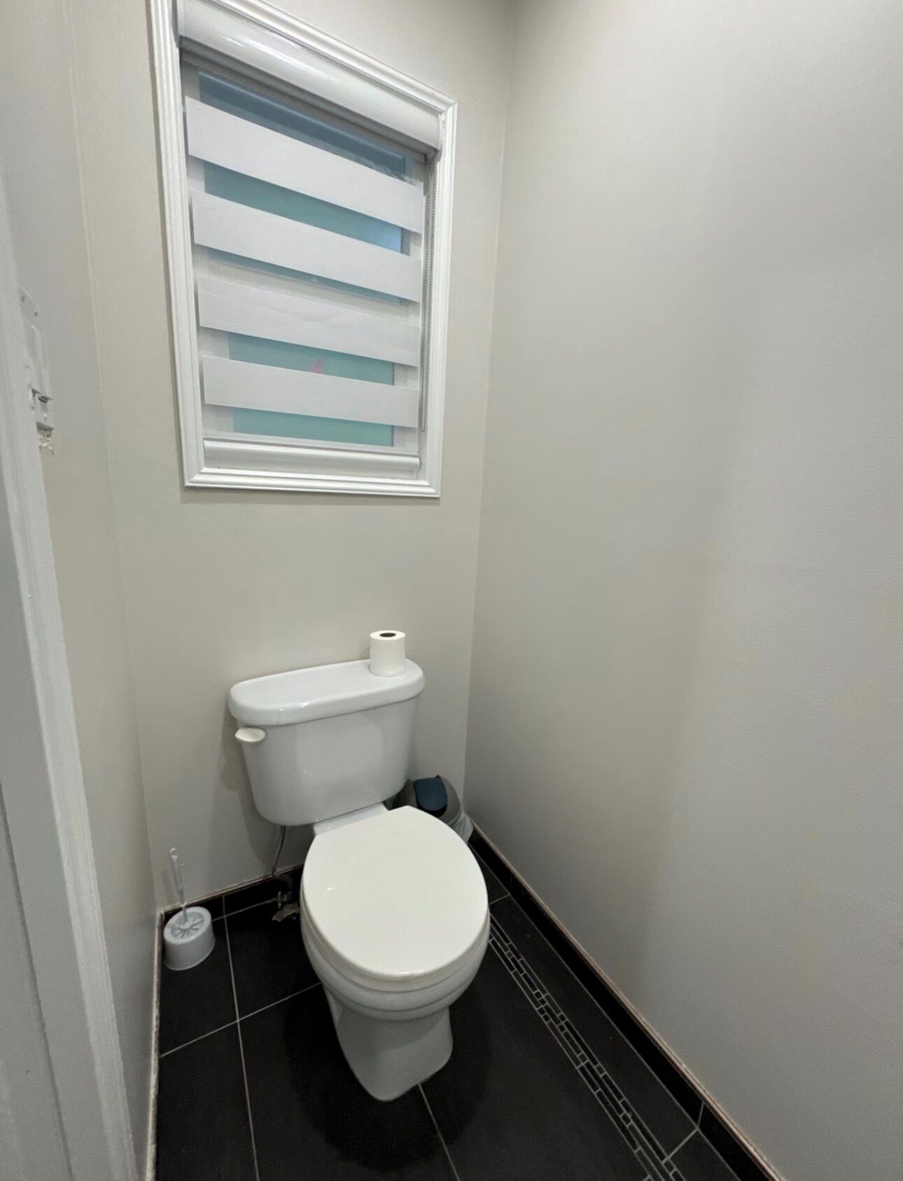 A small, clean bathroom with a toilet, a window with blinds, and a black tiled floor.