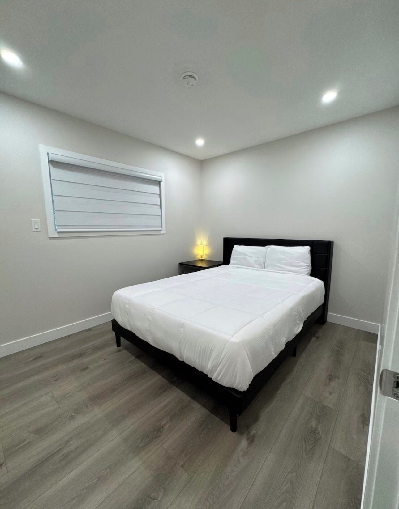 Une chambre à coucher comprenant un lit, une table de chevet, une lampe fixée au mur et une fenêtre avec des stores.