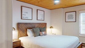 Quarto com cabeceira de madeira, cama com roupa de cama branca, duas mesas de cabeceira com luminárias e três frases emolduradas na parede.