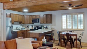Cozinha moderna com armários de madeira, eletrodomésticos de aço inoxidável e uma ilha central. Há uma área de jantar com mesa e cadeiras, e uma sala de estar com sofá e mesa de centro.