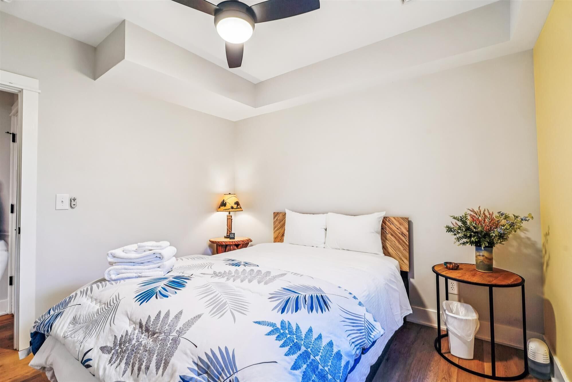 Un dormitorio con una cama, una mesita de noche con una lámpara, una pequeña mesa redonda con un florero y un cubo de basura.