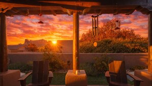 Un patio al aire libre con dos sillas de madera, un móvil de viento colgante y vistas a un atardecer sobre una cordillera.