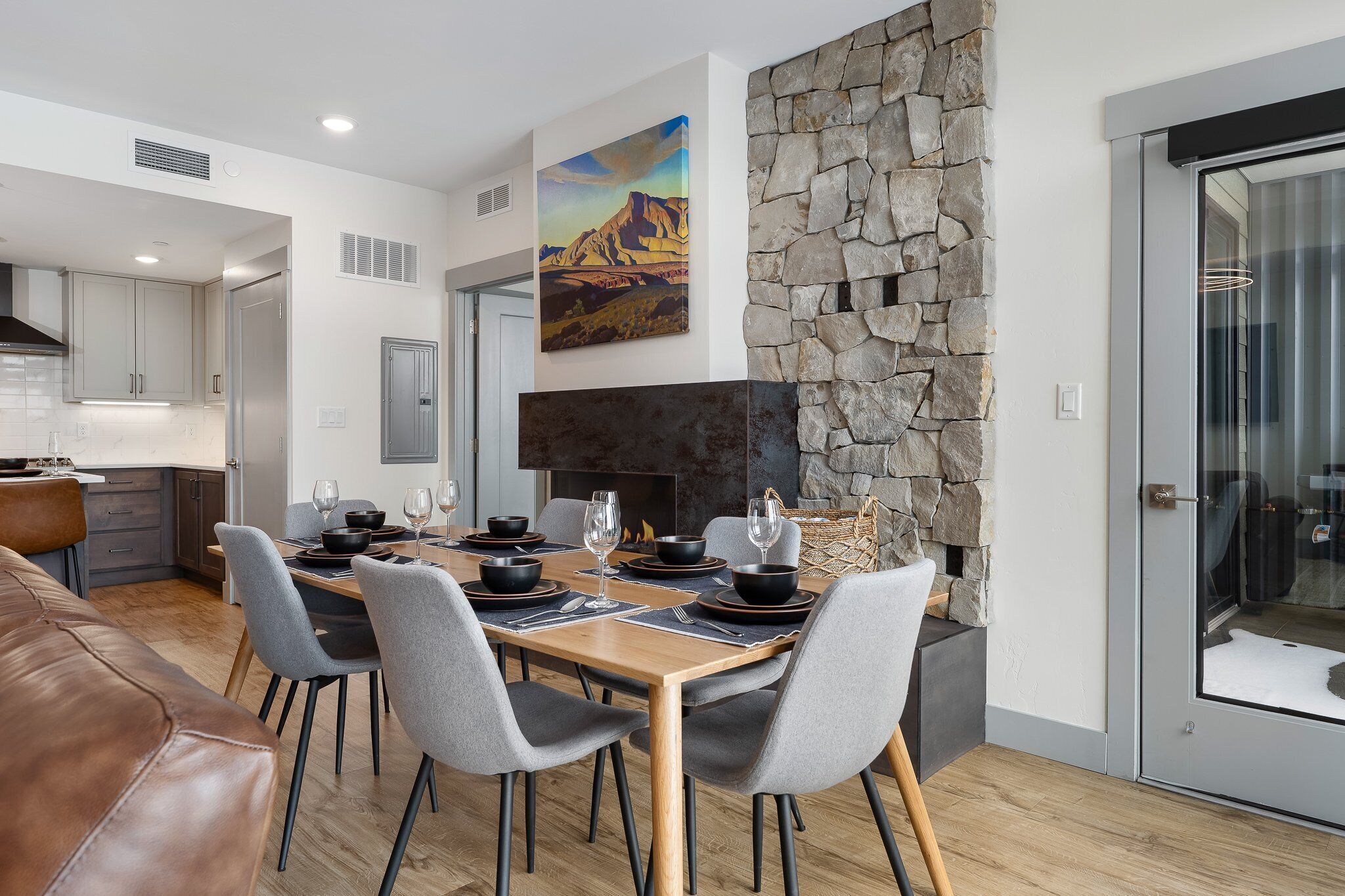 Un comedor moderno con chimenea de piedra, un cuadro de un paisaje montañoso y una puerta de vidrio que da a un balcón.