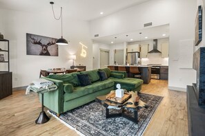 Ein modernes Wohnzimmer mit einem grünen Sofa, einem Couchtisch und einem Teppich. Ein Essbereich mit Tisch und Stühlen ist ebenfalls vorhanden. Im Hintergrund ist eine Küche mit modernen Geräten erkennbar.