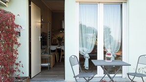 A small patio with a table and chairs, a window with curtains, and a wooden planter with red foliage.