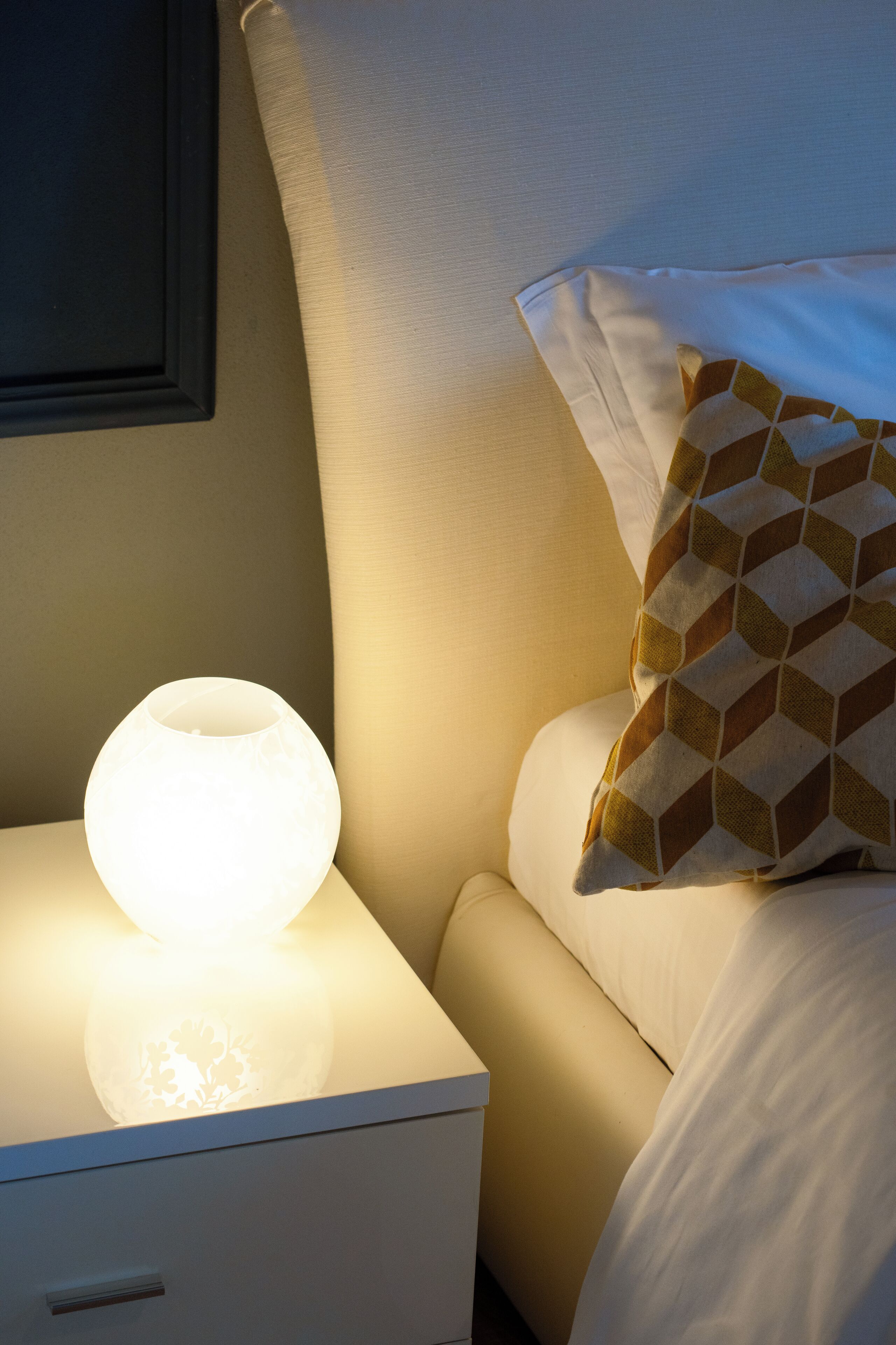 A neatly made bed with a patterned pillow and a lit bedside lamp on a white nightstand.