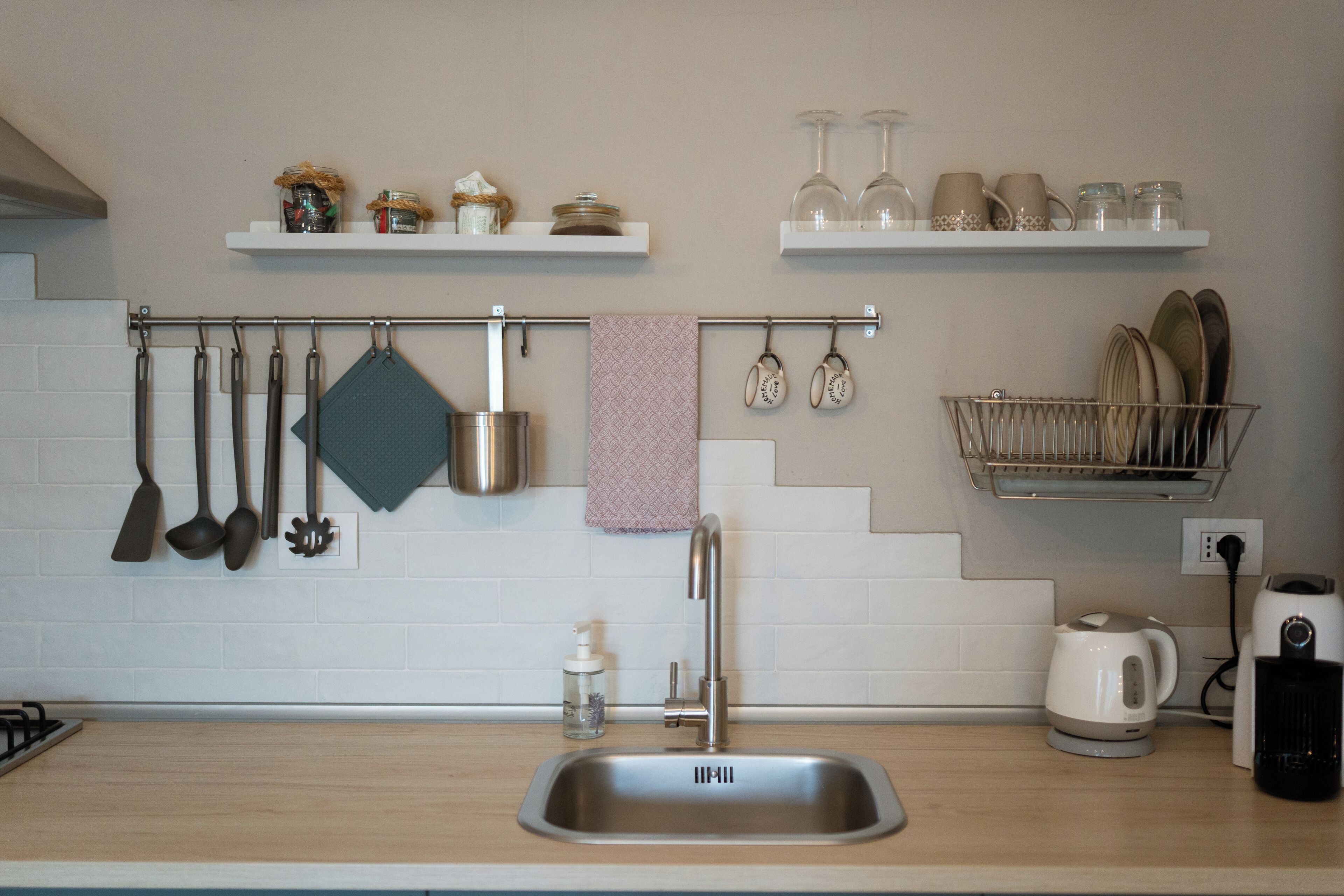 A kitchen with a stainless steel sink, hanging utensils, a dish rack, and a kettle.