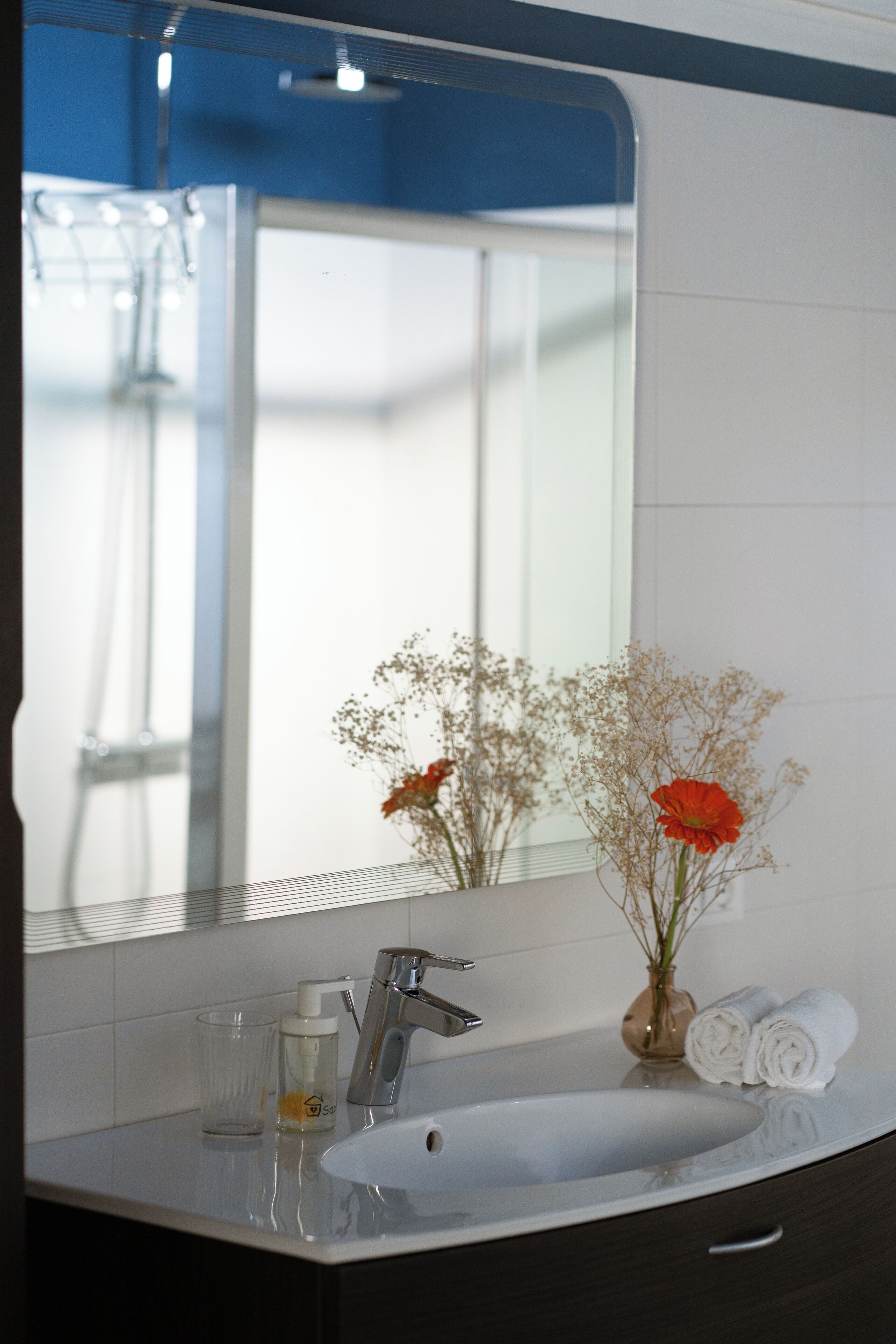 Un baño con un lavamanos blanco, un espejo, una toalla y un florero con flores.