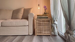 A cozy living room with a beige sofa, a wooden side table with books, a hanging light fixture, and a window with curtains. - WellHome Asti (Asti)