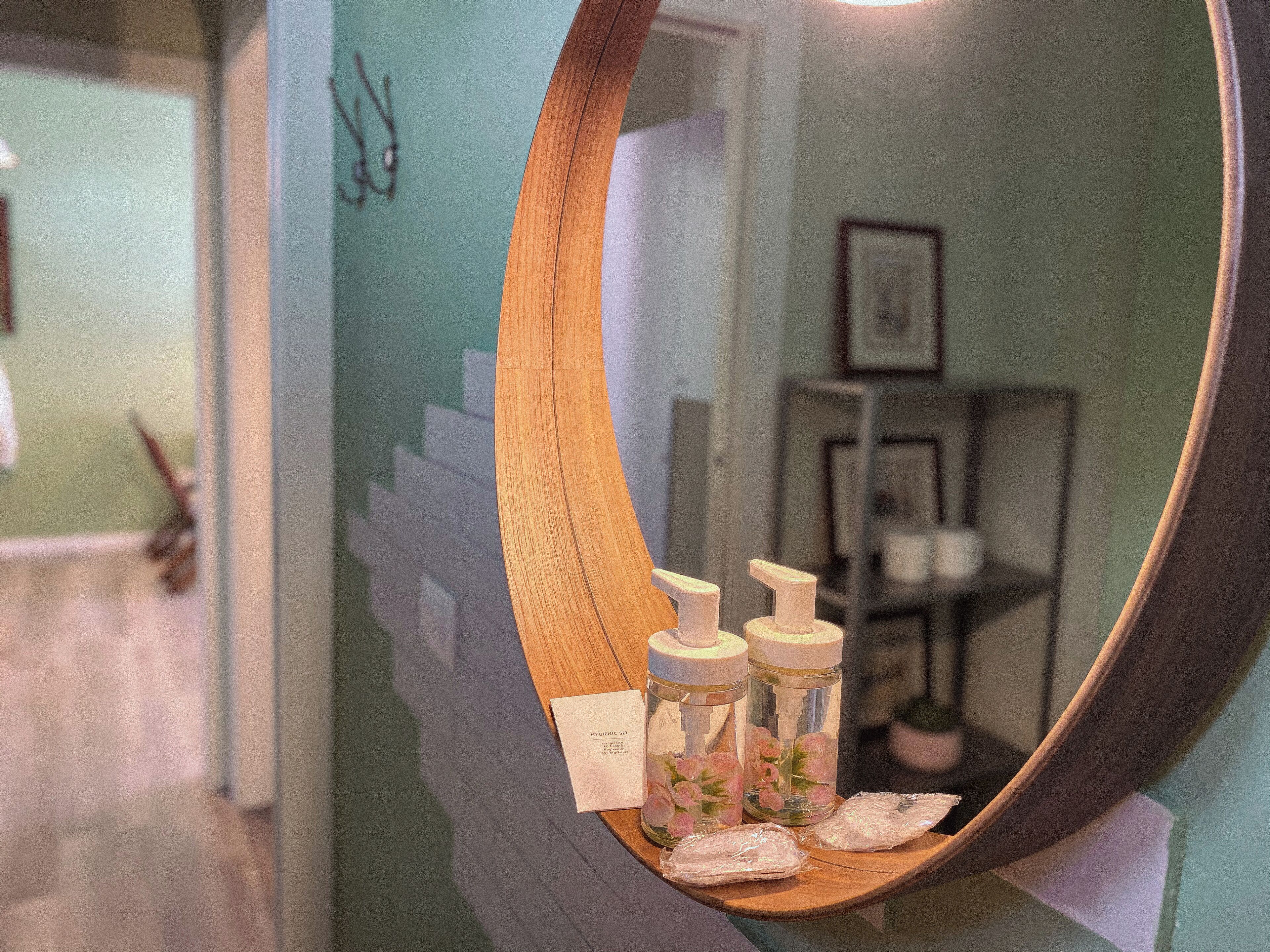 A mirror with a wooden frame reflecting a bathroom with a wooden shelf holding toiletries.