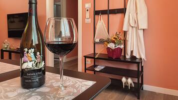 Une bouteille de vin Barbera d’Asti et un verre de vin rouge sont posés sur une table dans une pièce dont le mur est de couleur pêche, avec un étagère en métal et une petite table sur laquelle se trouve un vase de fleurs.