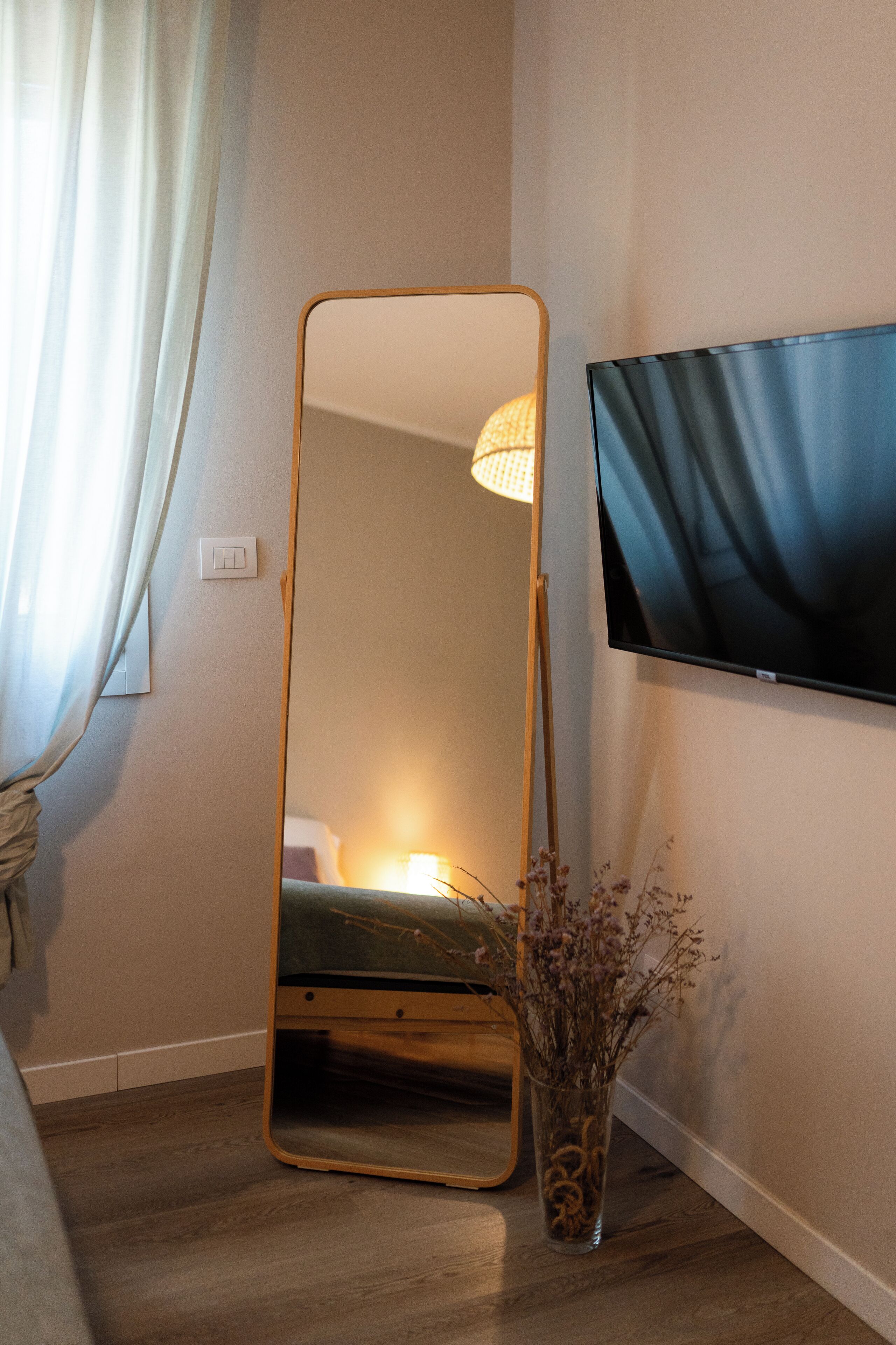 Ein Zimmer mit einem großen Spiegel, einem Flachbildfernseher, Holzboden, einem Bett und einer Vase mit getrockneten Blumen.