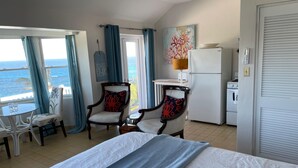 A hotel room with a bed, two armchairs, a small table, a refrigerator, and a view of the ocean.