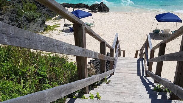 A wooden staircase leading to a beach with blue umbrellas and chairs.