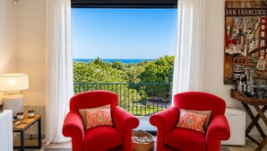 Une pièce avec deux fauteuils rouges, une vue sur l’océan, un tableau représentant San Francisco et une table avec des bouteilles.