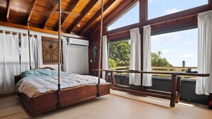 Un dormitorio con una cama de madera suspendida del techo, ventanas grandes con cortinas y un balcón con vistas a zonas verdes.
