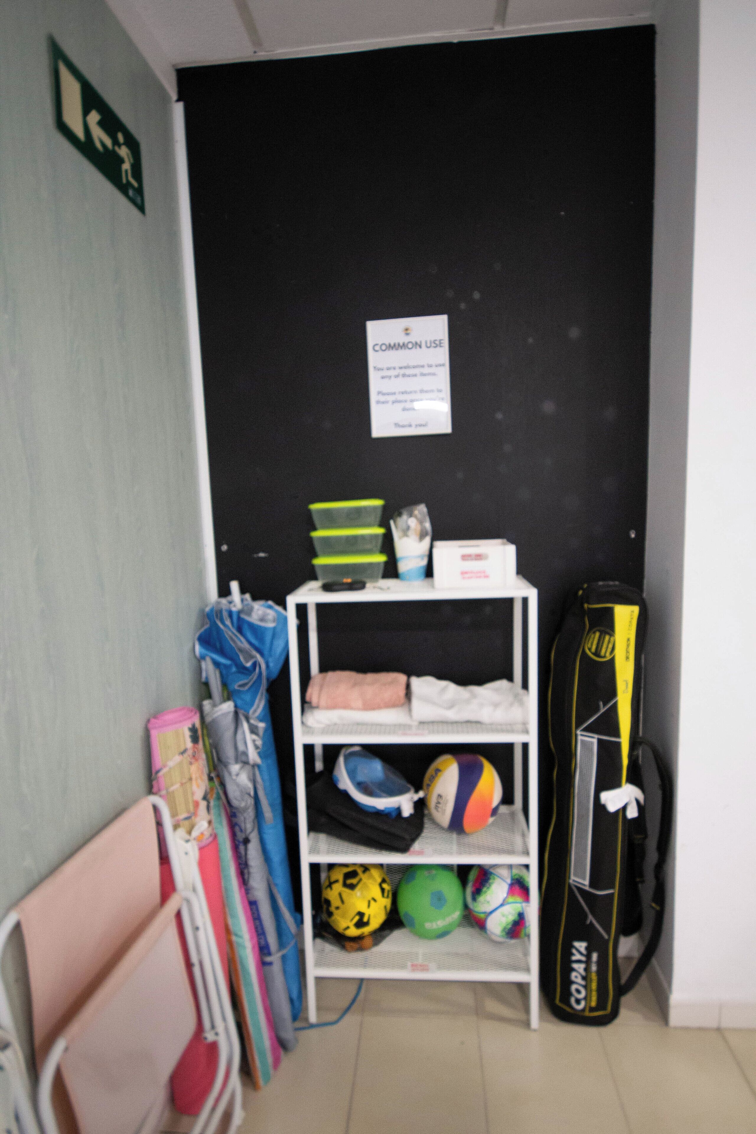 Un área de almacenamiento con estantes que contienen equipo deportivo, una pizarra con un cartel y una puerta con señal de salida de emergencia.