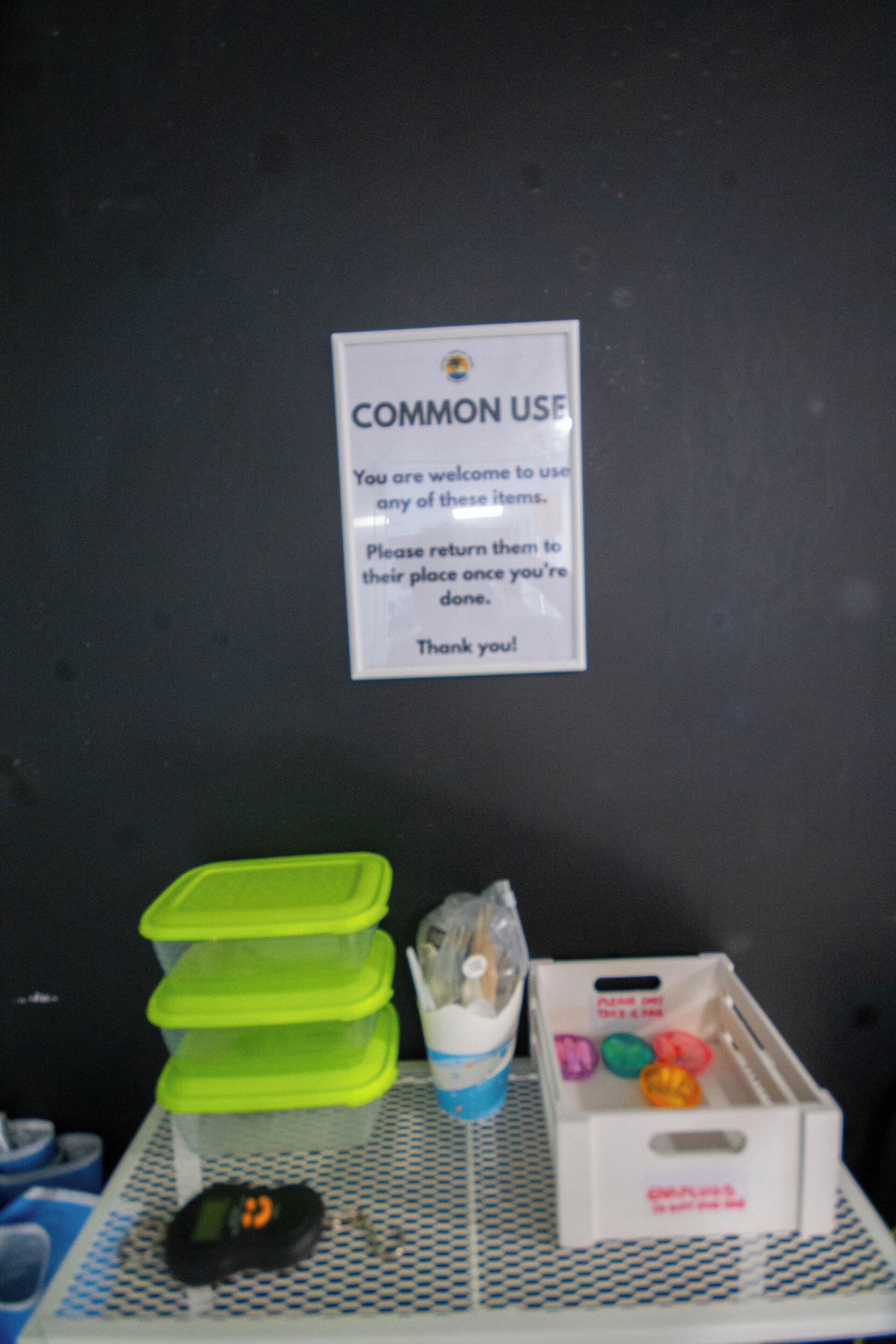 Um sinal na parede com a inscrição 'USO COMUM' e instruções para devolução de objetos. Abaixo do sinal, há recipientes de plástico, um saco pequeno e uma caixa com artigos coloridos.