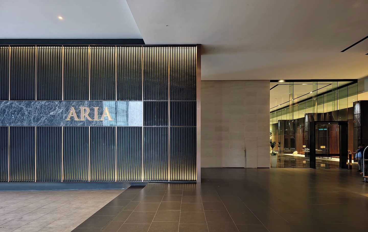 Un hall d’entrée moderne, orné d’un grand panneau « ARIA », un espace spacieux doté d’un sol carrelé et une entrée en verre donnant sur un espace détente.