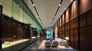 Un moderno vestíbulo de hotel con suelo de mármol, paredes de vidrio y paneles de madera. Hay zonas de estar con mesas y lámparas.
