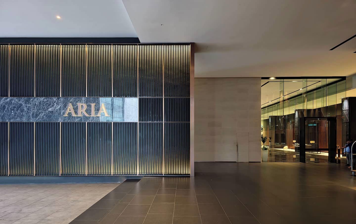 Eine moderne Lobby mit einem großen 'ARIA'-Schild, einem geräumigen Bereich mit Fliesenboden und einem Glastürdurchgang zu einer Sitzgelegenheit.