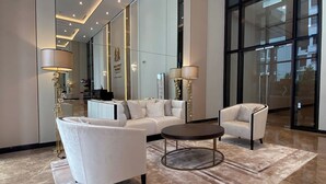 Eine großzügige Lobby mit einer weißen Sofagarnitur, zwei Sesseln, einem Couchtisch und hohen Stehlampen. Große Fenster lassen natürliches Licht herein und bieten Ausblick nach draußen.