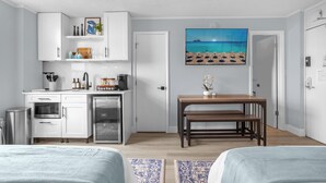 Una habitación de hotel compacta con kitchenette, un comedor con una mesa y banco de madera, y un cuadro grande con motivos playeros en la pared.
