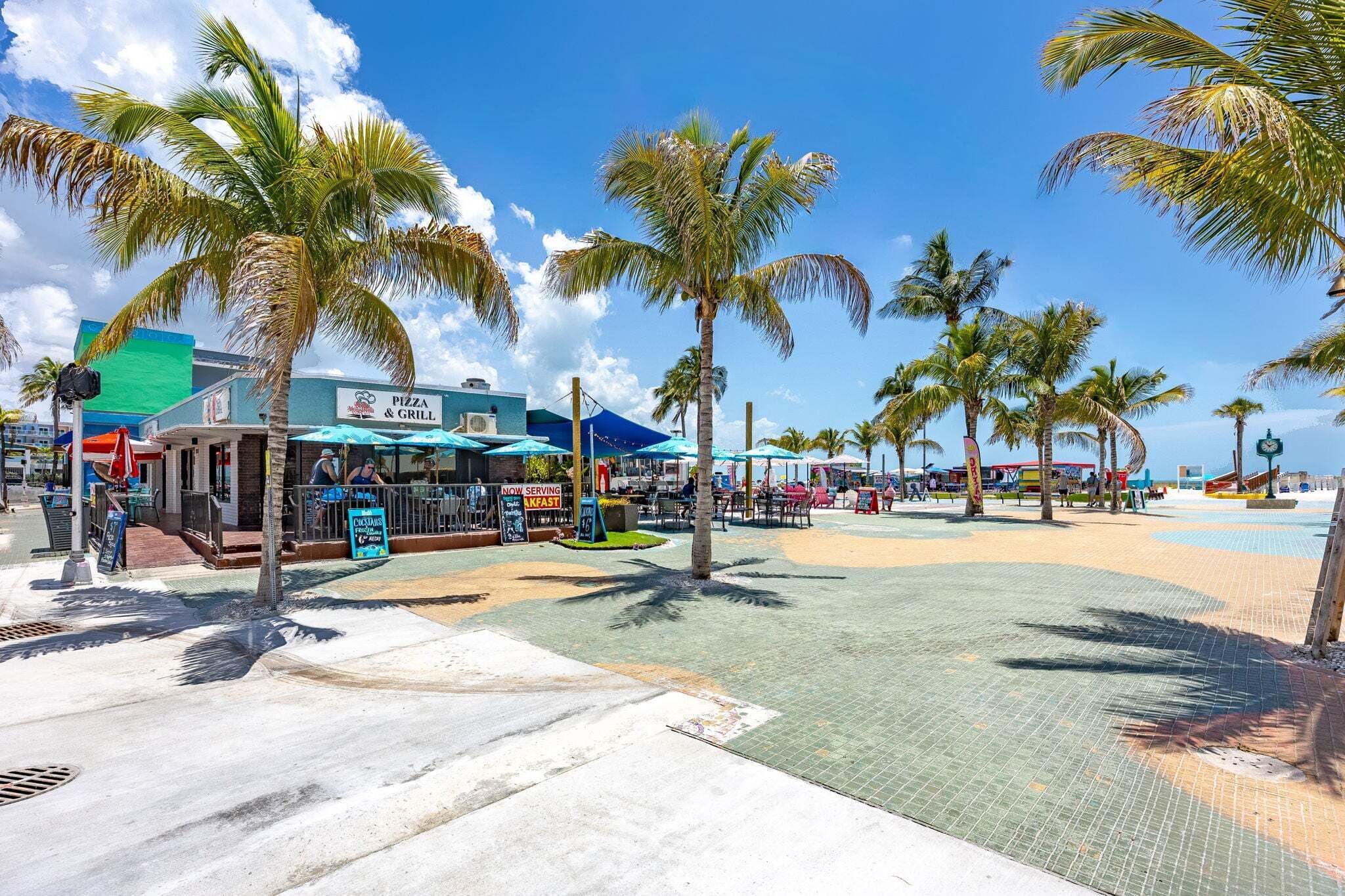 Une zone aménagée au bord de la plage, avec un restaurant servant des pizzas et des plats grillés, des palmiers et une promenade dallée.
