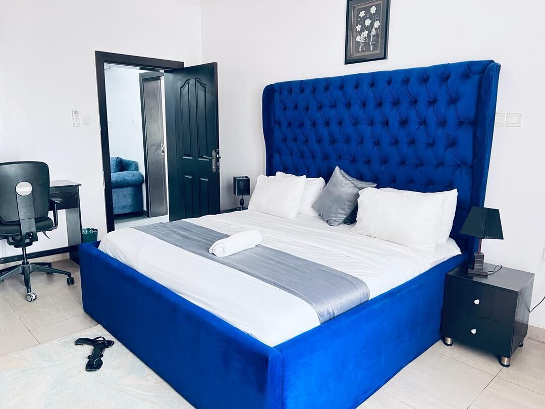A neatly made bed with a blue headboard, a small bedside table with a lamp, and a chair in a room with white walls and a door.