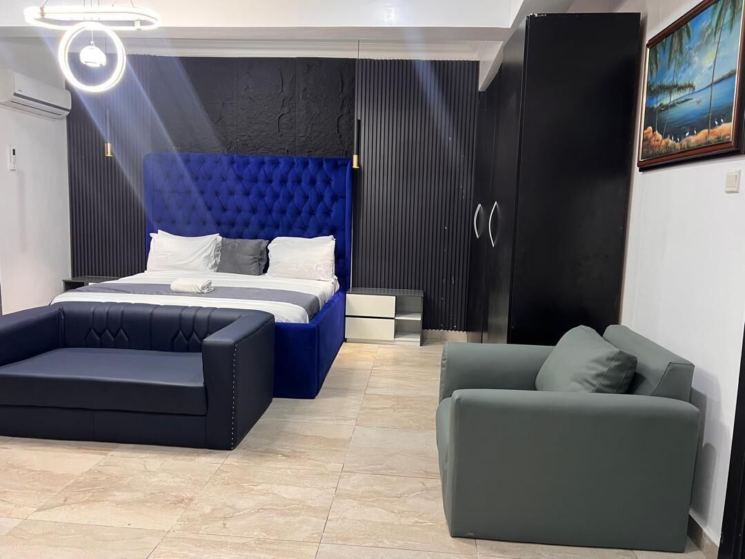 A modern bedroom with a tufted blue headboard, a large bed, a grey sofa, and a wardrobe.