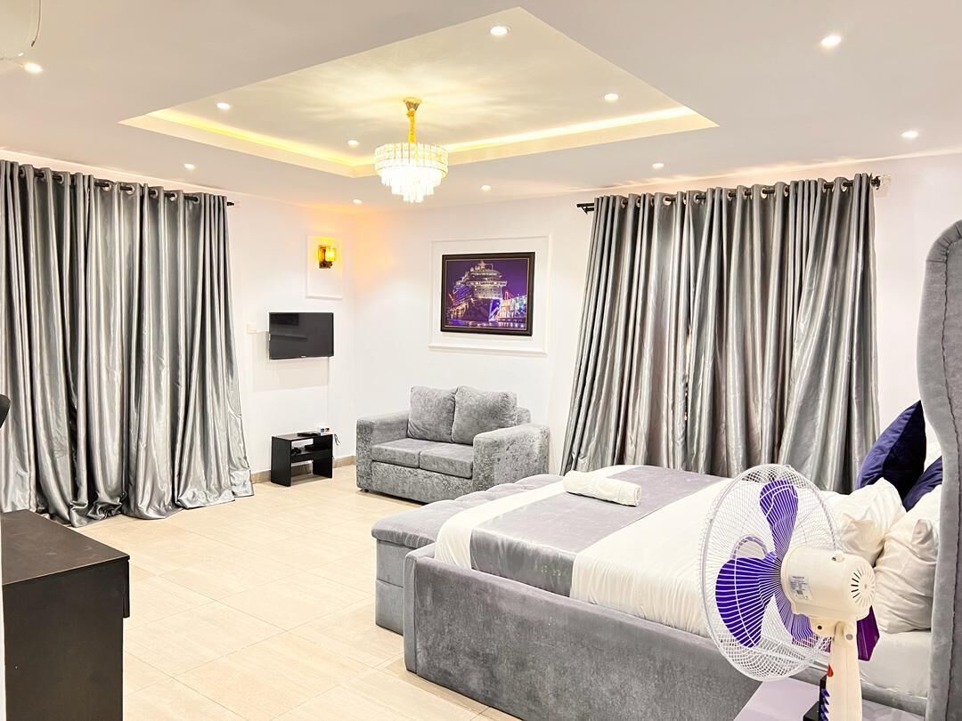 A modern bedroom with a large bed, a grey sofa, a standing fan, and a TV.