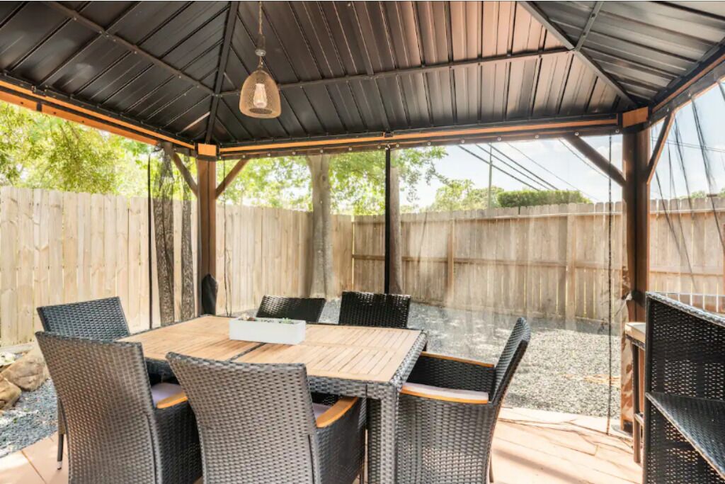 A covered patio with a wooden table and wicker chairs, surrounded by a wooden fence.