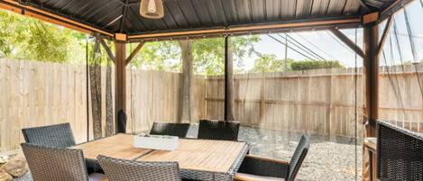 A covered patio with a wooden table and wicker chairs, surrounded by a wooden fence.