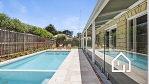 Ein rechteckiger Pool mit Fliesenrand, umgeben von einem Holzzaun und einer überdachten Terrasse mit Liegestühlen.