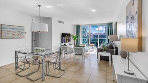 Un salon moderne avec une table à manger en verre, des canapés blancs et une grande fenêtre donnant sur des immeubles.