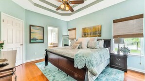 A bedroom with a large bed, a ceiling fan, a window with blinds, and a door.