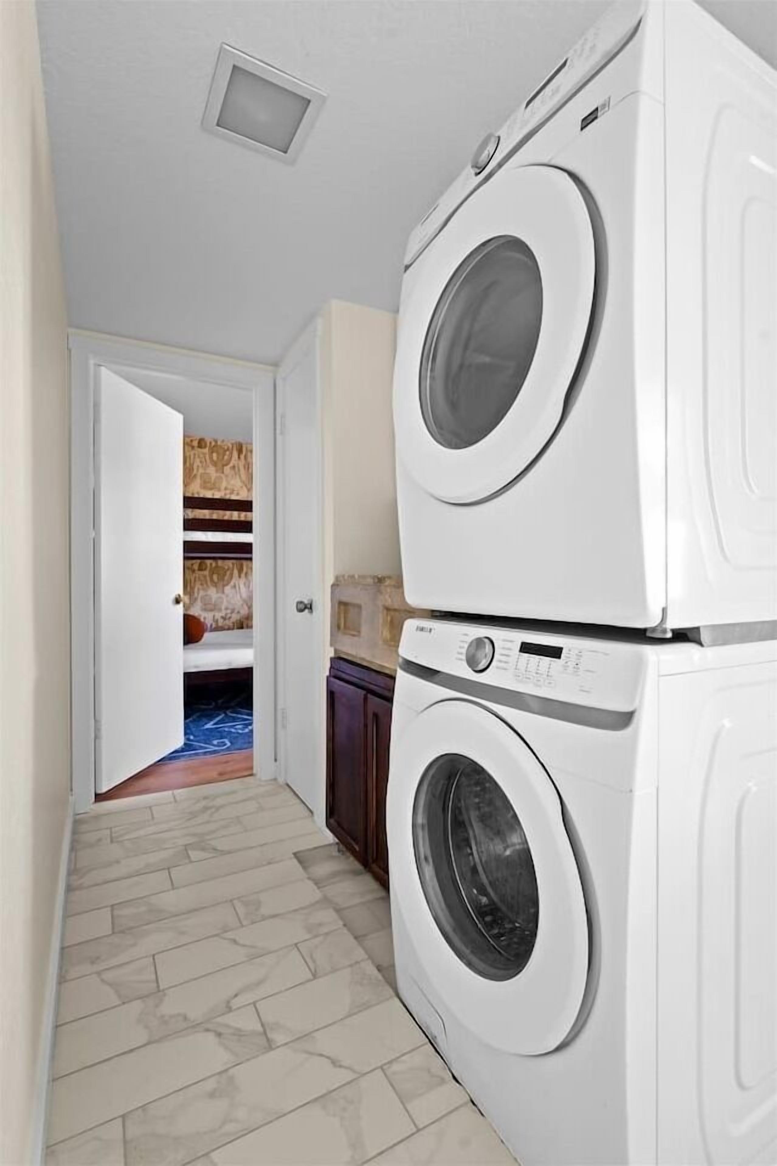 A laundry room with stacked washing machines and a door leading to a bedroom with patterned wallpaper.