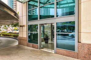 A modern building entrance with glass doors and windows, a potted plant, and a parking area visible through the glass. - Blueground | Burj Residence, pool, nr Burj Khalifa (Dubai)