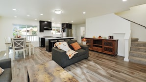 Ein modernes Wohnzimmer mit grauem Sofa, Holztisch und einem Essbereich mit weißem Tisch und Stühlen. Im Hintergrund ist eine Küche mit dunklen Schränken und einer weißen Arbeitsplatte zu sehen.
