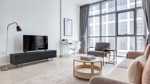 Ein modernes Wohnzimmer mit einem Flachbildfernseher auf einem schwarzen TV-Schrank, einem runden Holztisch, einem grauen Sofa und einem großen Fenster mit Vorhängen.