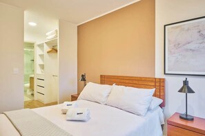 A hotel room with a bed, a nightstand, a lamp, a framed artwork on the wall, and a bathroom visible through an open door.