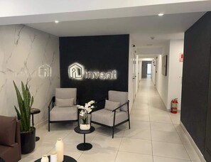 Eine moderne Lobby mit einer Rezeption, zwei Sesseln, einer Zimmerpflanze und einem an der Wand befestigten Schild mit der Aufschrift 'Invent'.