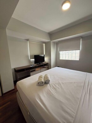 Ein Hotelzimmer mit Bett, Fernseher auf einem Tisch, einem Fenster mit Jalousien und einer Deckenleuchte.