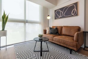 Ein modernes Wohnzimmer mit einem braunen Ledersofa, einem Teppich mit geometrischem Muster, einem kleinen runden Tisch mit Pflanze und einem großen abstrakten Gemälde an der Wand.